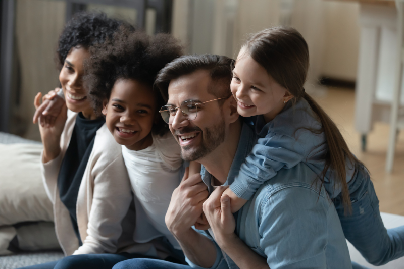 Close up multiracial family having fun at home together, smiling African American mother and Caucasian father piggy backing laughing little daughters, hugging and cuddling, enjoying leisure time