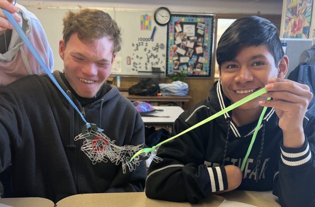 Two high school boys sit in a crowded classroom, laughing as they work on an experiment using magnets and ribbons to hold a pile of paperclips in the air between them.