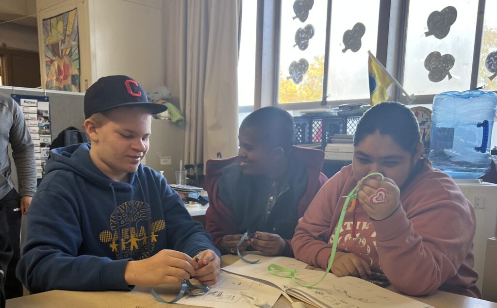 Three high school students in a special education class smile and sit at a classroom table, working on a science project using ribbons and magnets.