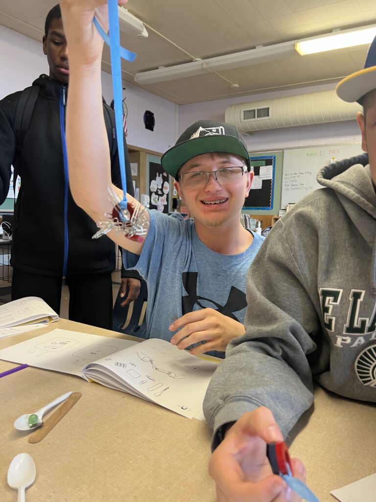 Seated between two classmates in a busy classroom, a male high school student with braces, a green baseball cap, and a blue tee shirt smiles for the camera while he holds a ribbon tied to a magnet that has picked up a handful of paper clips.