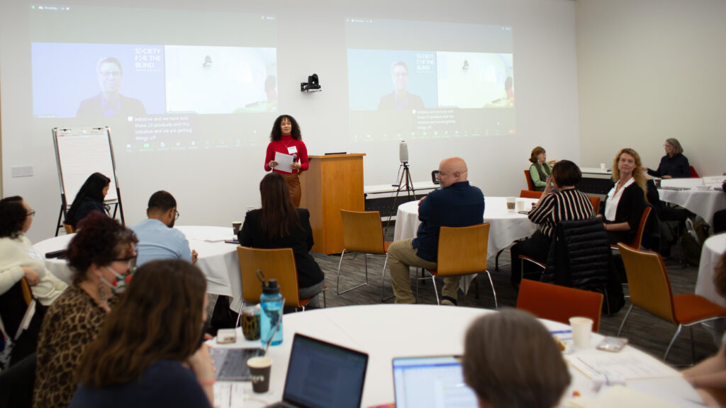Alyah Thomas, a biracial woman with long dark hair and a red turtleneck, presents to a seated crowd of Ability Central grantees in a modern event space.