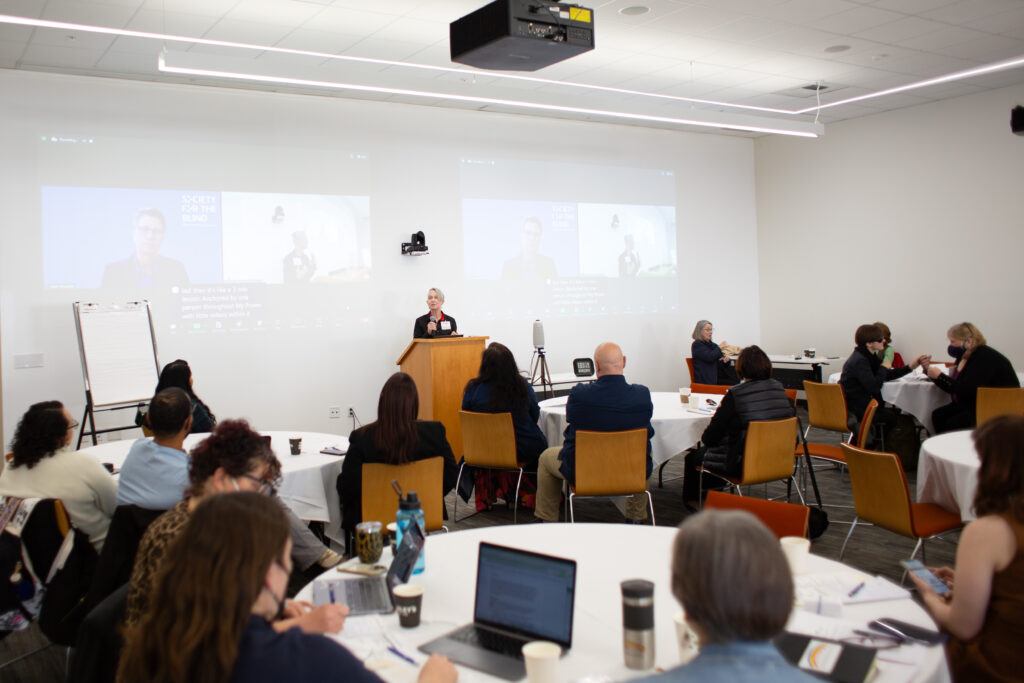 Erika Leonard, a white woman with short gray hair, presents to  the 2024 grantee gathering from the front of a modern event space. 